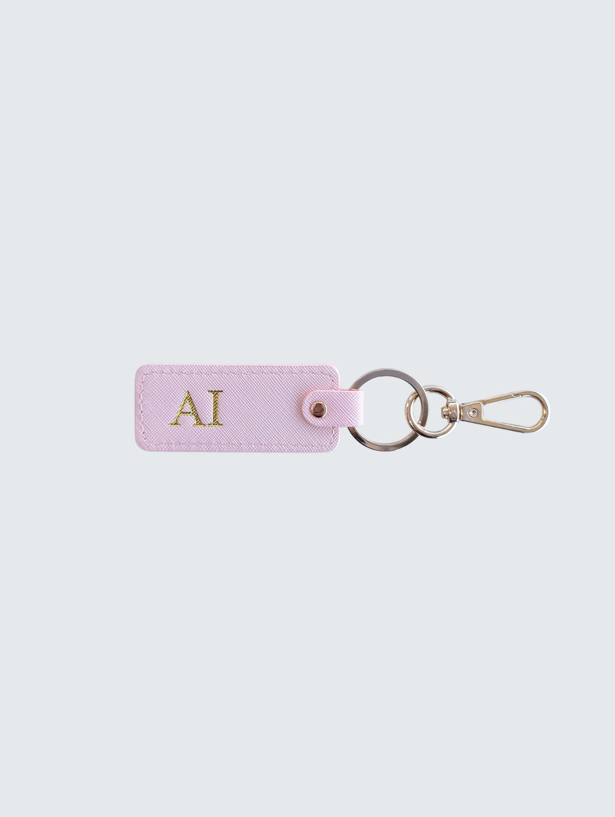 A light pink leather keychain tag with gold initials 'AI' monogrammed on the front, attached to a split keyring and gold-tone clasp, set against a solid light gray background.