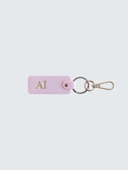 A light pink leather keychain tag with gold initials 'AI' monogrammed on the front, attached to a split keyring and gold-tone clasp, set against a solid light gray background.