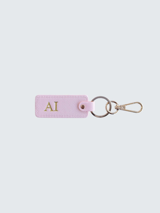 A light pink leather keychain tag with gold initials 'AI' monogrammed on the front, attached to a split keyring and gold-tone clasp, set against a solid light gray background.