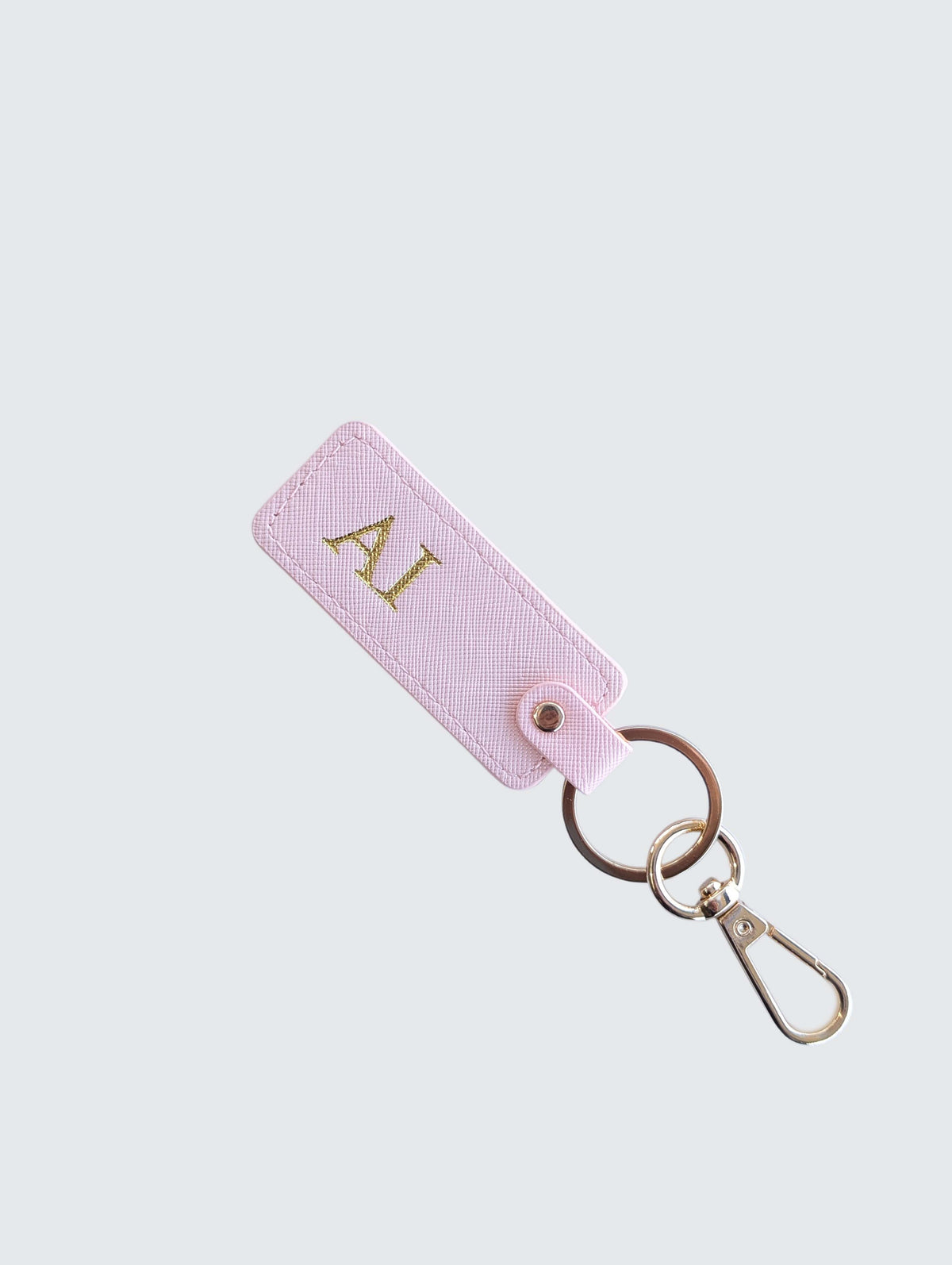 A light pink leather keychain tag with gold initials 'AI' monogrammed on the front, attached to a split keyring and gold-tone clasp, set against a solid light gray background.